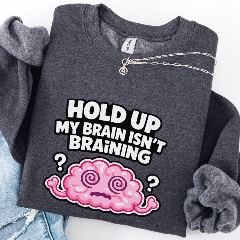 May include: A dark grey sweatshirt featuring the text "HOLD UP MY BRAIN ISN'T BRAINING" in white and black. A pink cartoon brain with swirly eyes and question marks is below the text. A silver necklace with a small pendant is on the sweatshirt.