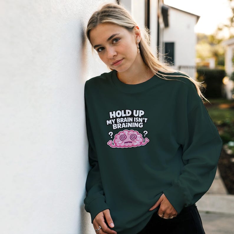 May include: Dark green sweatshirt featuring the phrase "HOLD UP MY BRAIN ISN'T BRAINING" and a cartoon brain graphic. The graphic is a pink brain with a confused expression and question marks. The sweatshirt is a casual style.