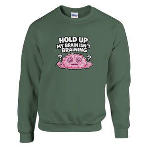 May include: A dark green sweatshirt featuring the text "HOLD UP MY BRAIN ISN'T BRAINING" and a cartoon brain. The brain is pink with swirling eyes and question marks on either side.