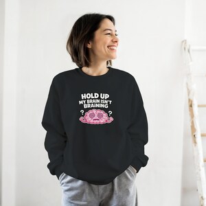 May include: Black crew neck sweatshirt featuring the phrase "HOLD UP MY BRAIN ISN'T BRAINING" above a pink cartoon brain with swirling eyes and question marks. The sweatshirt is a casual style, suitable for everyday wear.