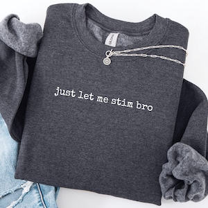 May include: A dark gray sweatshirt with the phrase "just let me stim bro" printed in white. A silver necklace with a circular pendant rests on the sweatshirt. The sweatshirt is folded with a pair of blue jeans visible.