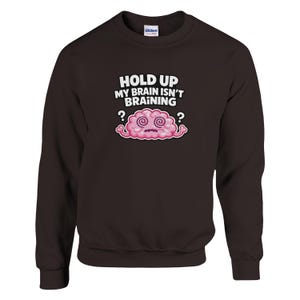 May include: A dark brown crew neck sweatshirt featuring the phrase "HOLD UP MY BRAIN ISN'T BRAINING" above a cartoon brain. The brain is pink with swirling eyes and question marks. The sweatshirt is a casual style.