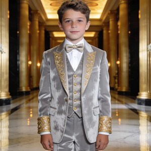 Puede incluir: Un traje de terciopelo plateado que incluye una chaqueta con solapas y puños bordados en oro, un chaleco a juego con botones dorados y una pajarita coordinada. El conjunto se lleva sobre una camisa blanca de cuello rígido, creando un estilo formal.