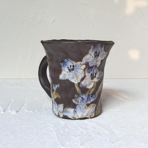 May include: A handmade ceramic mug with a dark brown exterior and a black handle. The mug features a floral design with blue and white flowers. This unique mug is ideal for tea or coffee. The mug is a unique piece of art.