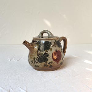 May include: A small, ceramic teapot with a lid, spout, and handle. The teapot is decorated with a pattern of green leaves and red fruit. The teapot is tan and brown in color.