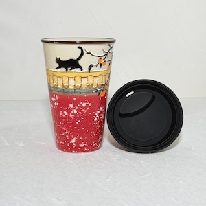 May include: A ceramic travel mug with a black lid. The mug features a black cat silhouette on a cream-colored background, with a yellow fence and red accents. The bottom half is red with white speckles.