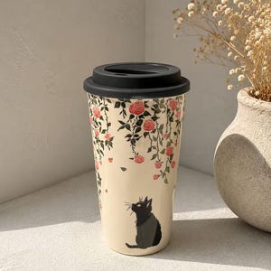 May include: A cream-colored travel mug with a black lid. The mug is decorated with pink roses and green leaves, and a black cat illustration. The mug is designed for hot or cold beverages.
