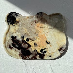 May include: A decorative tray in the shape of a cat's head. The tray is made of a stone material with a mix of colors, including cream, black, and brown. The surface has a polished finish and a unique pattern.