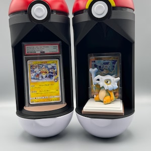 Può includere: Due espositori verticali bianchi e neri a forma di Poké Ball rosse e bianche. La teca sinistra contiene una carta Pokémon giapponese del 2018 graduata PSA con Pikachu. La teca destra contiene una statuetta di Cubone davanti a una carta.