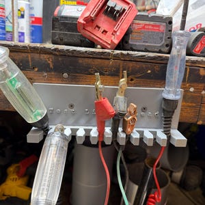 May include: A gray tool holder with multiple slots, holding various electrical testing tools. The tools include clear plastic voltage testers, red and black wires, and metal connectors. The text "Test Lead" is visible on the holder.