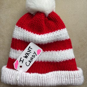 May include: A red and white striped knit hat with a white pom-pom on top. A tag attached to the hat reads "I WANT CANDY" in black marker. The hat has a white brim.