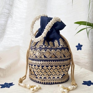 Puede incluir: Un bolso azul marino con cordón, bordado dorado intrincado y detalles de lentejuelas. Cuenta con un asa de cuentas blancas y borlas. El diseño incluye patrones florales y geométricos, creando un accesorio elegante.