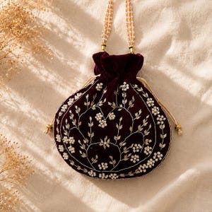 May include: A deep burgundy velvet drawstring pouch featuring intricate white beadwork and metallic thread embroidery in a floral pattern. The bag includes a multi-strand pearl handle and gold-toned metal accents at the drawstring closure points.