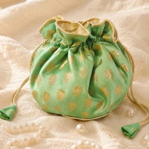 May include: A green drawstring pouch with gold polka dots and gold trim. The pouch is made of a silky fabric and has a drawstring closure with gold tassels. Pearls are scattered around the pouch.