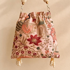 Puede incluir: Un bolso tipo saco de color rosa claro con cierre de cordón, que presenta un intrincado bordado de elefantes, motivos florales y patrones arquitectónicos en tonos rojo, verde y dorado. Incluye herrajes metálicos dorados y cuentas blancas.