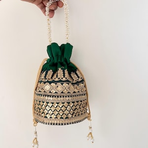Puede incluir: Un bolso verde oscuro con cordón, bordados dorados y lentejuelas. El bolso tiene un asa de perlas y un cordón dorado. La parte inferior del bolso tiene borlas doradas con perlas. El bolso está sostenido por una mano.