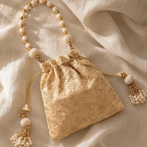 May include: A beige drawstring pouch featuring intricate gold floral embroidery. The bag includes a handle made of white beads and two side drawstrings, each finished with a decorative tassel composed of small white beads and gold accents.