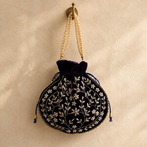 Puede incluir: Un bolso tipo saco de terciopelo morado oscuro con cierre de cordón, adornado con intrincados bordados plateados y pequeñas perlas blancas. Incluye un asa hecha de múltiples hilos de perlas blancas y cordones azules con borlas metálicas doradas.