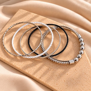 Puede incluir: Un conjunto de cinco pulseras apiladas con diversos diseños, incluyendo una banda plateada texturizada, una banda de cuerda blanca trenzada, una banda plateada fina, una banda de cuentas negras y una banda plateada con esferas y un muelle.