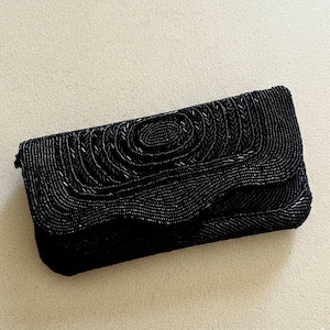 May include: A black beaded clutch bag with a flap. The front of the bag has a spiral design, and it is covered in small, dark beads. The rectangular shape is perfect for carrying essentials.