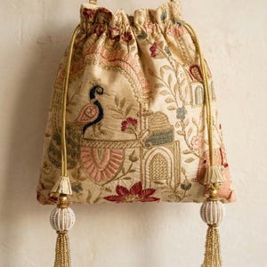 May include: Cream-coloured drawstring bag with intricate embroidery featuring a peacock, floral, and architectural design. The bag has gold-coloured drawstrings and tassels with white beads and gold fringe.