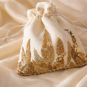 May include: Cream-coloured drawstring pouch with gold beaded and sequined floral design. The pouch is made of a soft fabric and features a delicate, ornate pattern. The bag is closed with a white drawstring.