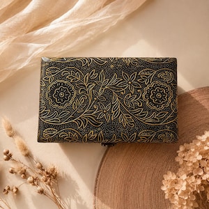 May include: A rectangular black box featuring an intricate gold-coloured floral and vine pattern etched across the lid. The design includes stylised blossoms and leaves with a fine stippled texture background, and a small metal latch at the front.