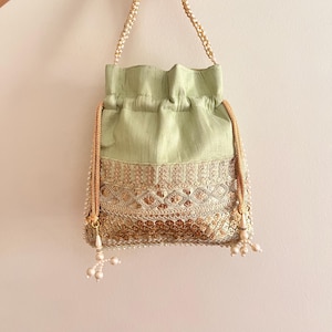 Puede incluir: Un bolso tipo saquito de color verde salvia claro con intrincados bordados dorados y detalles de lentejuelas en la parte inferior. El bolso incluye un asa de cuentas de perlas y cordones dorados con puntas de perla para el cierre.