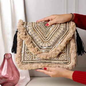 May include: Beige clutch with intricate beadwork and sequin details. The flap features a geometric pattern, and the edges are adorned with fringe. Black tassels hang from the sides, adding a touch of elegance to this stylish accessory.
