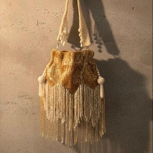 Puede incluir: Un bolso de mano dorado con cuentas, borde festoneado y largas borlas. El bolso tiene una correa color crema y detalles de borlas. El bolso cuelga contra una pared de color neutro.