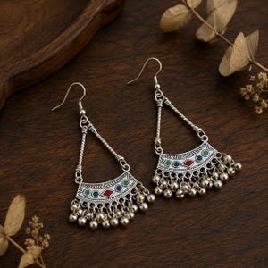 May include: A pair of silver-toned dangle earrings featuring a triangular frame and a curved base plate. The base plate is decorated with small inlaid red, blue, and green enamel shapes and is finished with a row of dangling silver-toned metal beads.