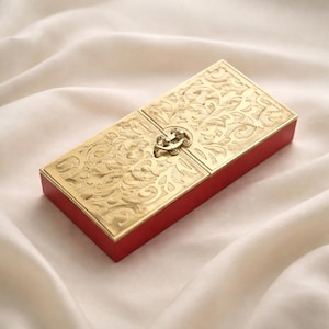 Puede incluir: Una caja rectangular con una base de color rojo vibrante y una tapa de metal dorado con un intrincado patrón floral en relieve. La tapa está asegurada por un mecanismo de cierre central de metal que une las dos mitades de la superficie.