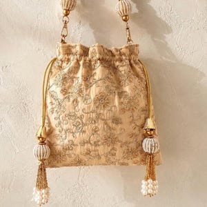 Peut inclure: Une pochette à cordon beige ornée de broderies florales dorées complexes. Le sac comprend une fermeture à cordon doré avec deux grands pompons décoratifs en perles blanches et accents dorés, ainsi qu'une anse perlée assortie avec fermoirs dorés.