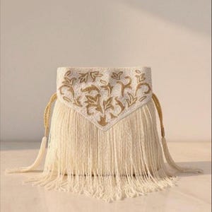 May include: Cream-coloured fringed handbag with gold embroidered floral design. The bag features a triangular flap with intricate gold embroidery and long, cream-coloured fringe. It has gold rope handles.