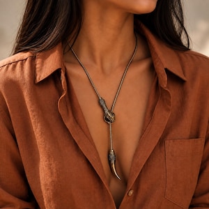 Op de afbeelding: Een metalen lariat-ketting met een gestructureerde slangenkopsluiting die de ketting op zijn plaats houdt, met een puntige, taps toelopende hanger aan het uiteinde. De ketting heeft een donkere, antieke messing afwerking en wordt over een roestkleurig overhemd gedragen.