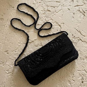 May include: A rectangular black clutch bag covered in intricate black beadwork arranged in a swirling, wave-like pattern. The bag features a matching black beaded shoulder strap and a flap closure that follows the curved edge of the design.