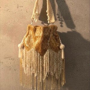 May include: A gold beaded drawstring handbag featuring a dense, shimmering surface of metallic gold beads. The bag is adorned with long, cascading cream-colored beaded fringe at the base and sides, complemented by two decorative beaded tassels.
