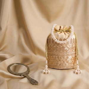 Puede incluir: Un bolso dorado con cordón, con intrincados detalles de lentejuelas y bordados, adornado con un asa de cuentas blancas y borlas. Un pequeño espejo de mano de estilo antiguo descansa cerca, sobre un fondo suave y neutro.