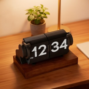 May include: A black retro flip clock displaying the time 12:34 PM, set on a wooden base. The clock has white numerals and is placed on a wooden surface, with a small potted plant and a desk lamp in the background.
