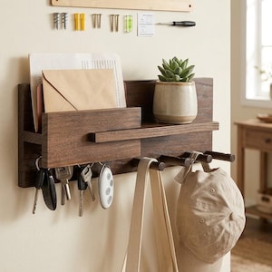 May include: A dark wood wall-mounted organizer with a shelf, key hooks, and hanging pegs. It holds keys, a plant in a ceramic pot, mail, a tote bag, and a baseball cap. Mounting hardware is included above the organizer.