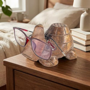 May include: A wooden turtle-shaped glasses holder with a pair of burgundy-framed glasses resting on it. The holder is brown and has carved details. The glasses have a rectangular shape.