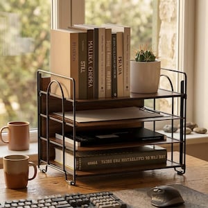 May include: A black metal desk organizer with three shelves, holding books, papers, and a small potted cactus. The books have titles like "PRADA" and "mayhem". Two ceramic mugs sit on the wooden desk, along with a keyboard and mouse.
