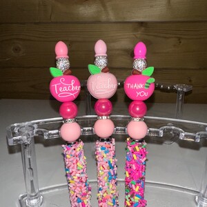 May include: Three decorative pens with pink and silver details. Each pen has a pink apple charm with the word "Teacher" or "Thank You". The pen barrels are covered in colourful sprinkles, and the pens are displayed in a clear stand.