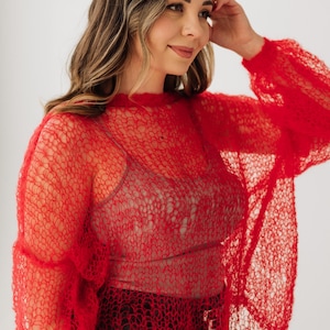 Könnte beinhalten: Ein roter, offen gewebter Pullover mit langen Ärmeln. Der Pullover besteht aus einem locker gestrickten Material, das einen durchsichtigen Effekt erzeugt. Das Model trägt ein neutralfarbenes Oberteil darunter. Der Hintergrund ist einfarbig weiß.