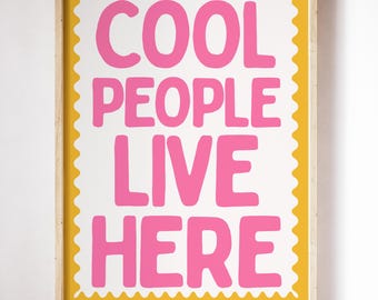 May include: A framed art print with the words "COOL PEOPLE LIVE HERE" in bright pink, set against a white background. The print has a yellow border with a scalloped edge and is encased in a light wood frame. The artwork is displayed on a white wall.