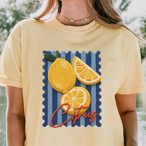 May include: Yellow t-shirt featuring a citrus-themed design. The design includes embroidered lemons and a slice, set against a blue and white striped background. The word "Citrus" is written in red cursive. The shirt is short-sleeved.