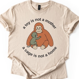 May include: Beige t-shirt featuring a cartoon orangutan embracing a smaller monkey. The text "a toy is not a mother, a cage is not a home" is printed around the image in black. The shirt has short sleeves and a crew neck.