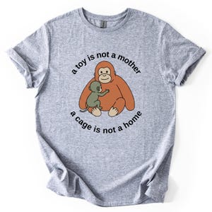May include: A heather gray t-shirt featuring a cartoon orangutan hugging a baby orangutan. The text "a toy is not a mother, a cage is not a home" is printed around the orangutans in a circular pattern.