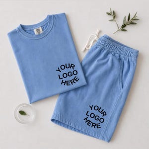 May include: A light blue t-shirt and matching shorts set featuring the black text "YOUR LOGO HERE" printed on the chest of the shirt and the lower leg of the shorts. The shorts have an elastic waistband with white drawstring cords.