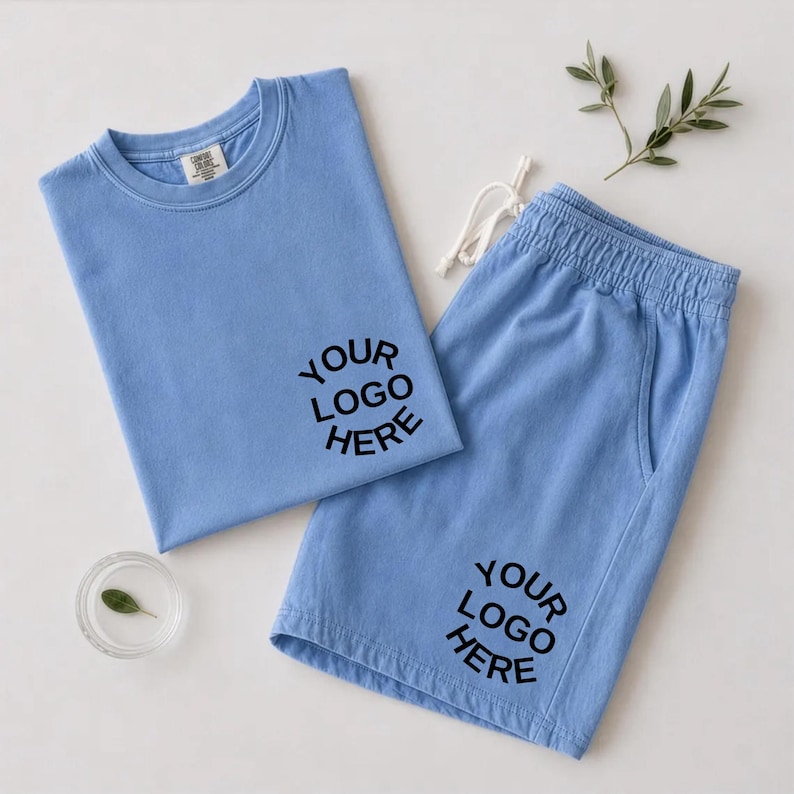 May include: A light blue t-shirt and matching shorts set featuring the black text "YOUR LOGO HERE" printed on the chest of the shirt and the lower leg of the shorts. The shorts have an elastic waistband with white drawstring cords.